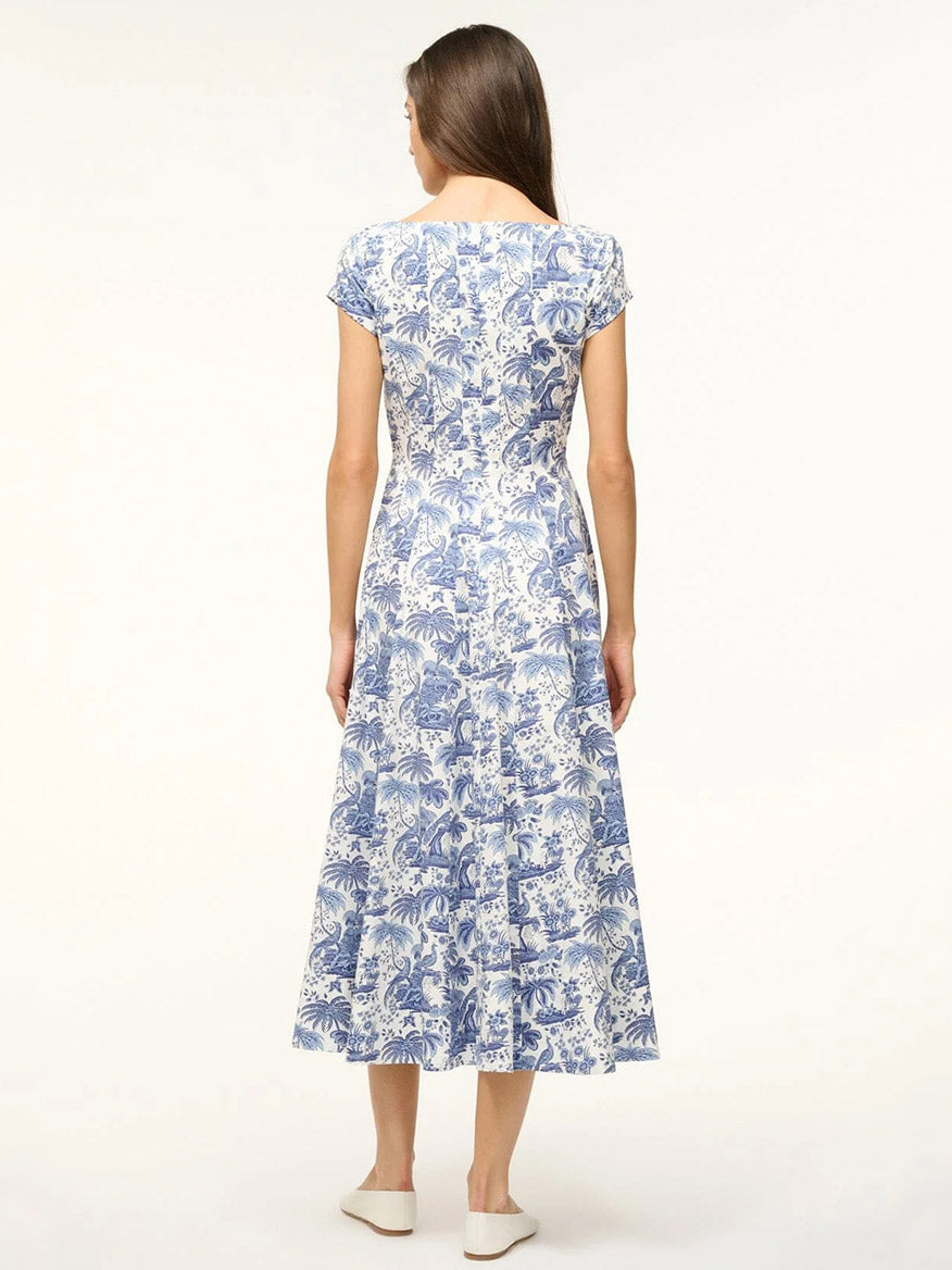 Woman stands with her back to the camera, wearing the STAUD Wells Short Sleeve Dress in Blue Toile with an a-line skirt and white shoes, set against a plain background.