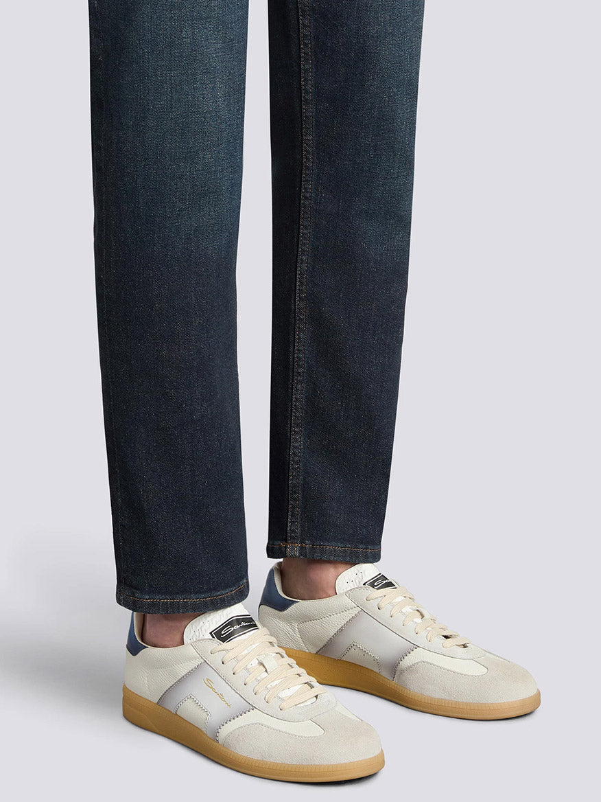 Person wearing dark blue jeans and Santoni Double Buckle Sneaker in White/Blue, standing against a plain light gray background.