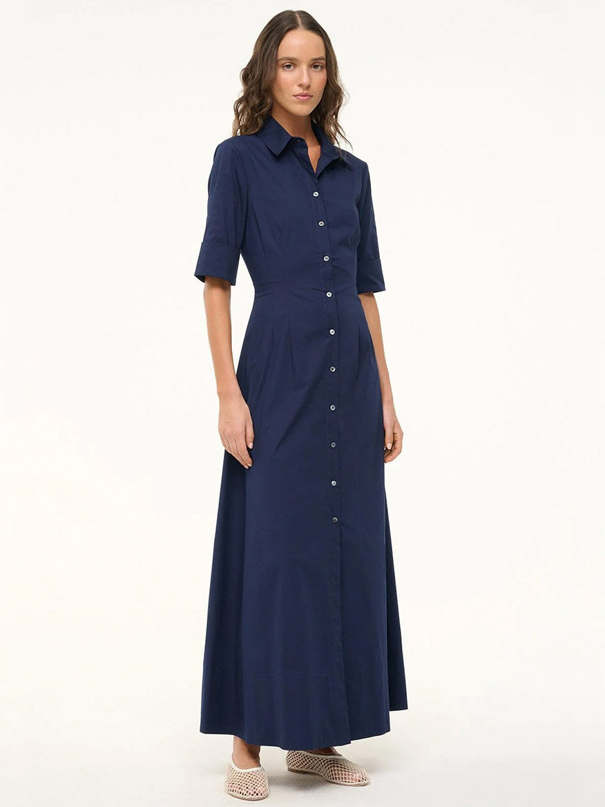 A woman stands in the STAUD Joan Maxi Dress in Navy, a stretchy cotton poplin maxi shirt dress with short sleeves and a collared button-down design, paired with light shoes against a plain background.