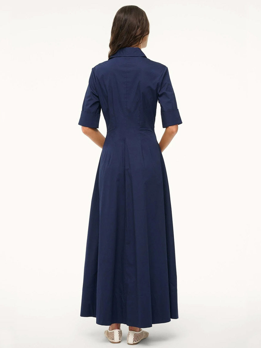 A woman with long brown hair and beige shoes faces away, wearing the STAUD Joan Maxi Dress in Navy—an ankle-length shirt dress made from stretchy cotton poplin.