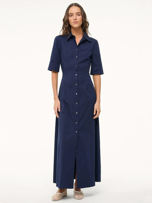 A woman faces forward in a STAUD Joan Maxi Dress in Navy—a button-down, collared, short-sleeve cotton poplin maxi shirt dress—and beige woven slip-on shoes against a plain light background.