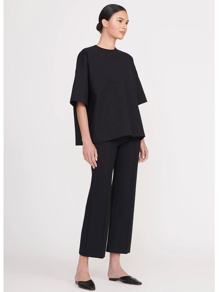 A woman stands against a plain background wearing a loose black top, the STAUD Knack Pant in Black (mid-rise ankle-length ponte knit), and black slip-on shoes.