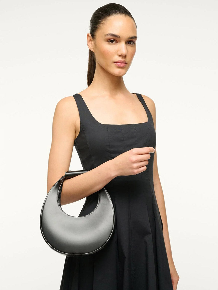 A woman in a black sleeveless dress holds the STAUD Mini Moon Bag in Black—a chic leather crescent bag—against a plain, light background.