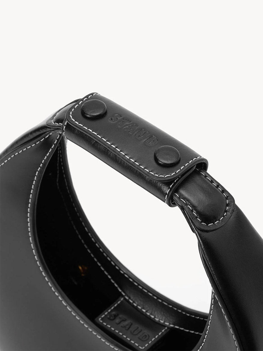 Close-up of the STAUD Mini Moon Bag in Black: features a black leather handle with white stitching, two black metal studs, an embossed "STAUD" logo, and a glimpse of its sleek crossbody strap.