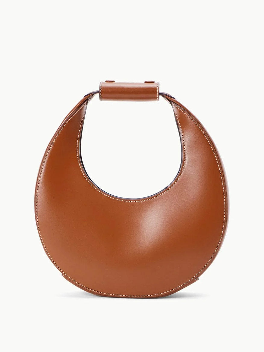 The STAUD Mini Moon Bag in Tan is a leather crescent bag with a structured handle, visible stitching, and a sleek silhouette, shown against a plain white background.