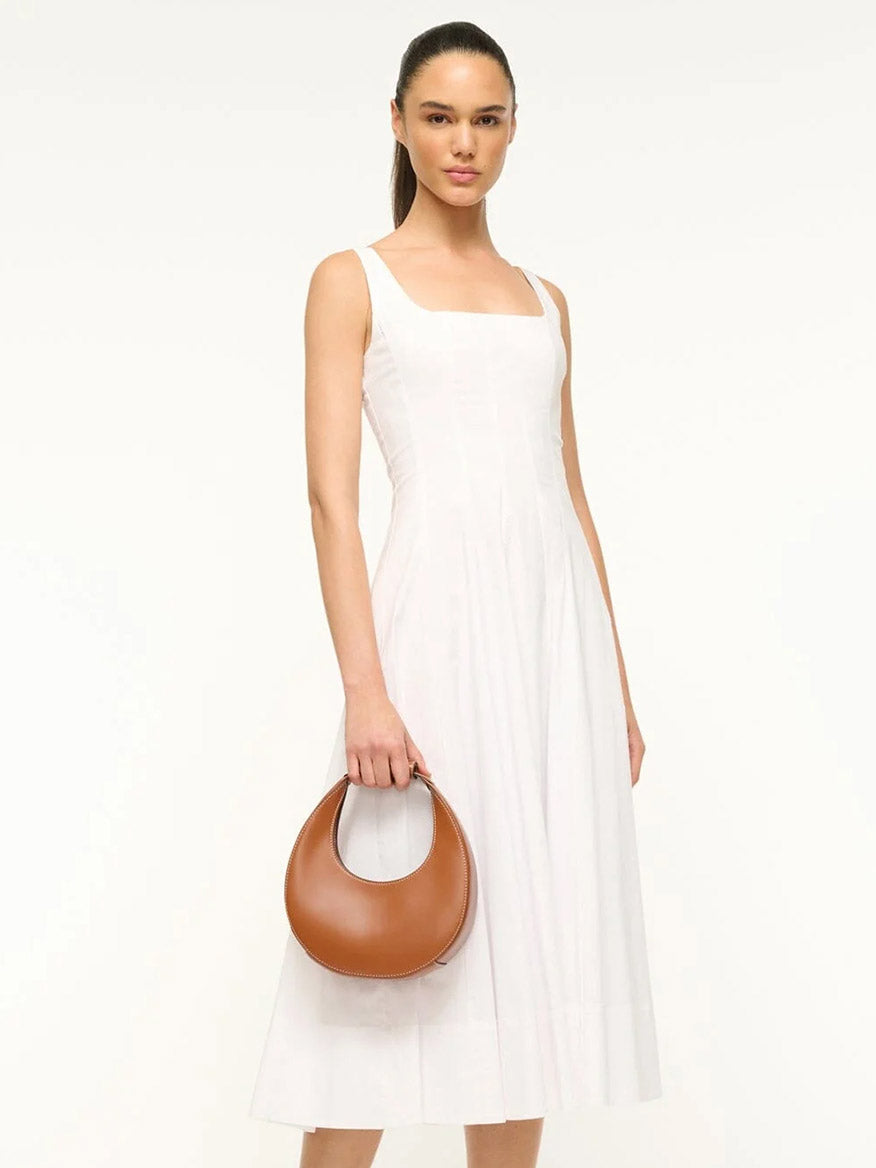 Woman wearing a sleeveless white midi dress, holding the STAUD Mini Moon Bag in Tan with a crossbody strap, standing against a plain white background.