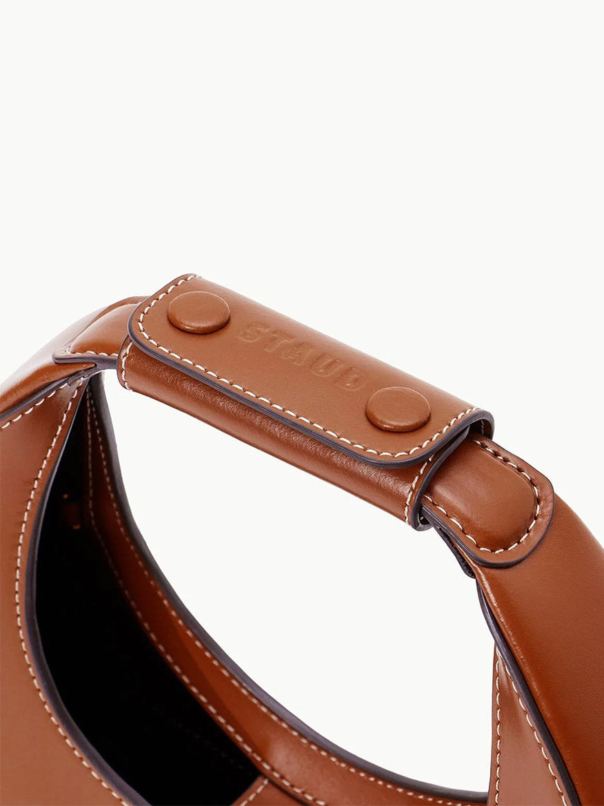 Close-up of the STAUD Mini Moon Bag in Tan, featuring a brown leather crescent handle with white stitching and an embossed leather brand label, showcasing its elegant details.