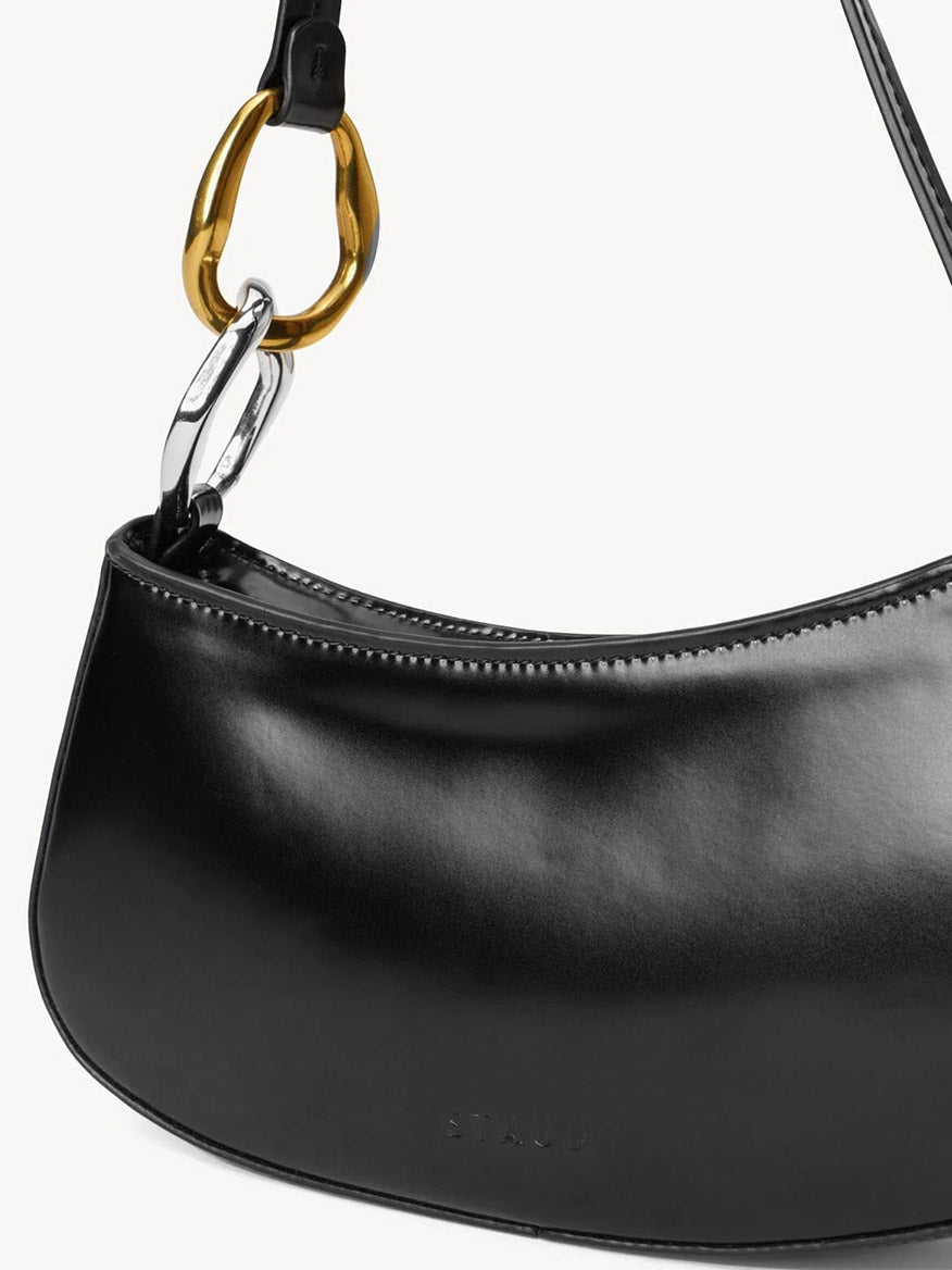 A close-up of the STAUD Ollie Bag in Black, a polished leather mini shoulder bag with gold and silver two-tone hardware ring accents near the strap attachment.
