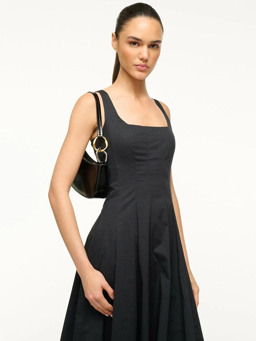 A woman in a sleeveless, square-neck black dress faces forward, holding the STAUD Ollie Bag in Black—a polished leather mini shoulder bag with two-tone hardware.
