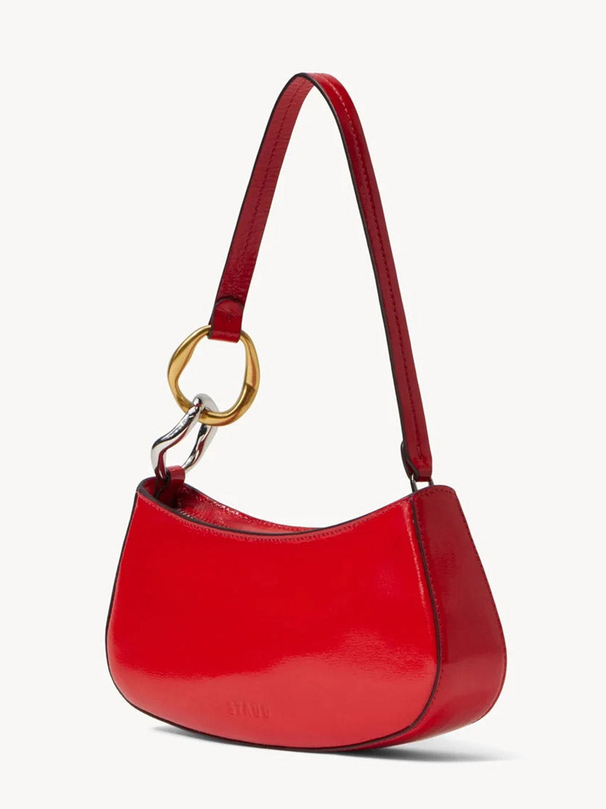 The STAUD Ollie Bag in Cherry is a small, shiny red polished leather mini shoulder bag featuring a curved shape, short strap, and gold-tone metal ring accent on one side.