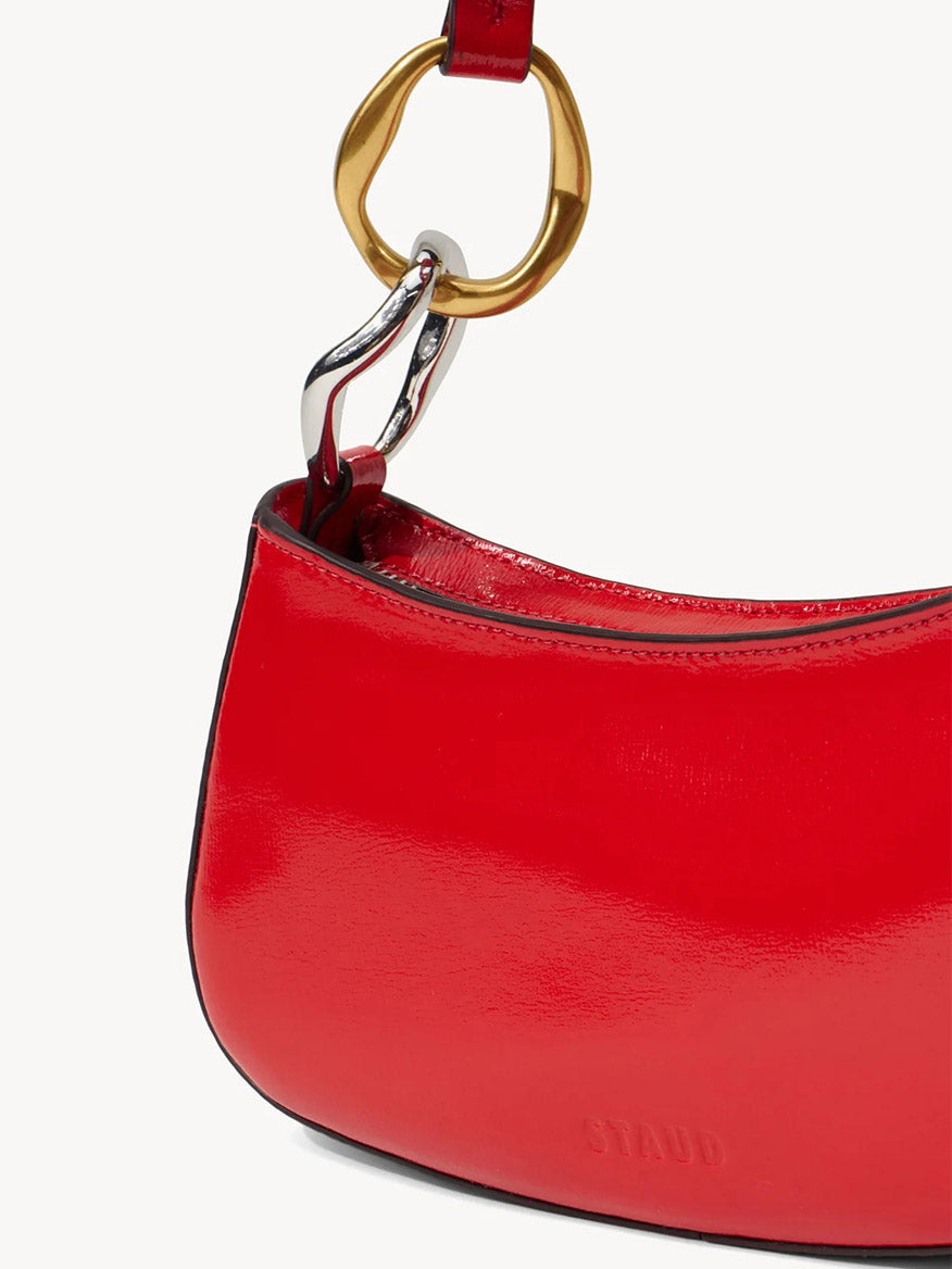 Close-up of the STAUD Ollie Bag in Cherry, featuring shiny red polished leather, gold and silver strap hardware, and "STAUD" embossed near the bottom.