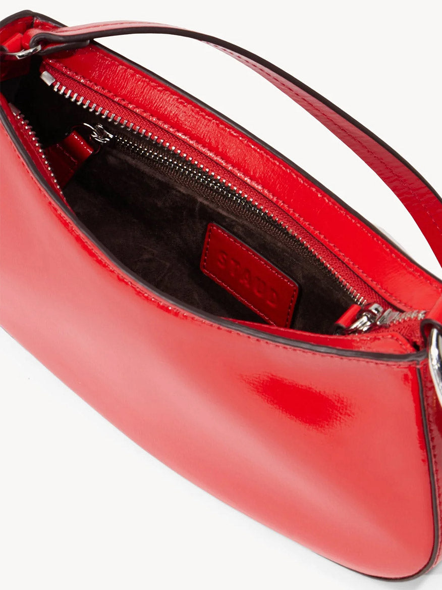 A close-up of the STAUD Ollie Bag in Cherry, an open glossy red leather shoulder bag with a zip closure, reveals a zipper pocket and the "STAUD" logo label inside.