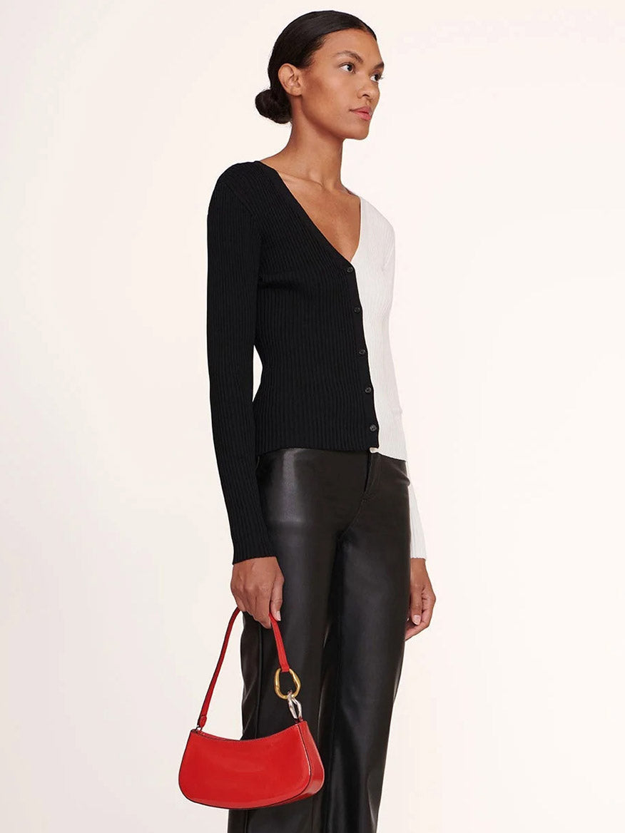 A woman wearing a black and white ribbed cardigan, black leather pants, and holding the STAUD Ollie Bag in Cherry stands against a plain background.