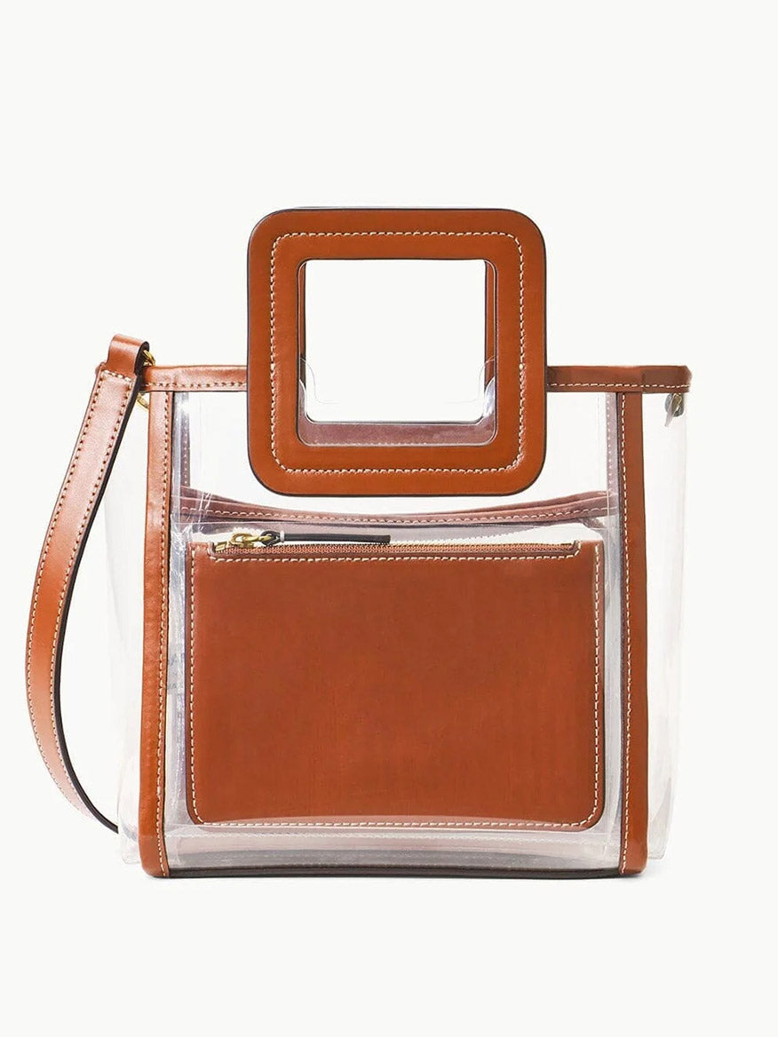 The STAUD Stadium Mini Shirley Bag in Clear/Tan features brown leather trim, a square handle, and a detachable strap. This stadium-approved PVC handbag includes a matching brown zippered pouch inside.