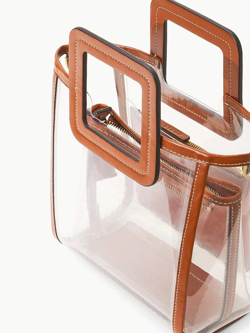 The STAUD Stadium Mini Shirley Bag in Clear/Tan features a clear PVC body, brown leather trim, square handles, and a zippered inner pouch—making it a chic, stadium-approved accessory showcased on a white background.