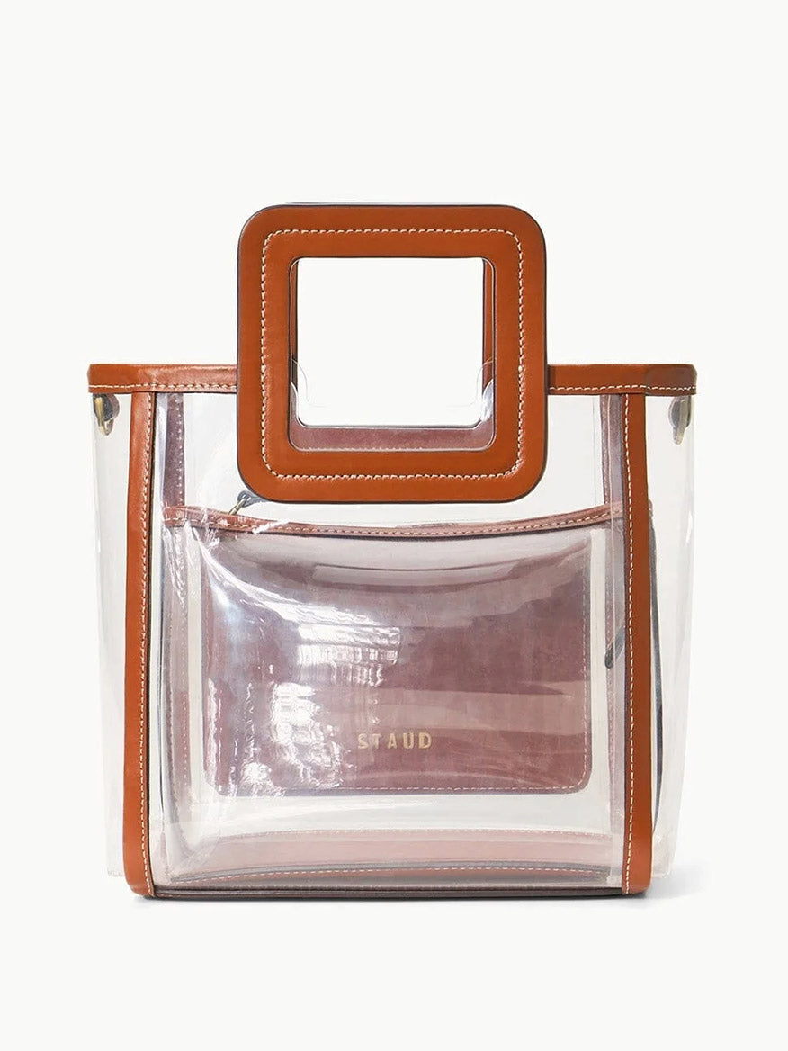 The STAUD Stadium Mini Shirley Bag in Clear/Tan is a stadium-approved transparent PVC tote with brown leather trim, square handles, and includes a matching clear zip pouch featuring "STAUD" in gold lettering.