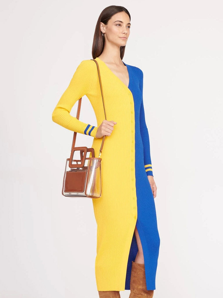 A woman in a long, buttoned yellow and blue ribbed dress carries the STAUD Stadium Mini Shirley Bag in Clear/Tan, a trendy clear PVC handbag with brown trim, paired with brown boots.