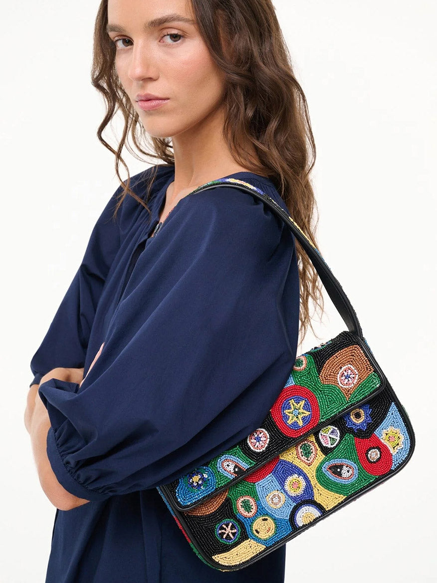 A person wearing a navy blue top holds the STAUD Tommy Beaded Bag in Black Millefiori, featuring colorful embroidered circular patterns, over their shoulder.