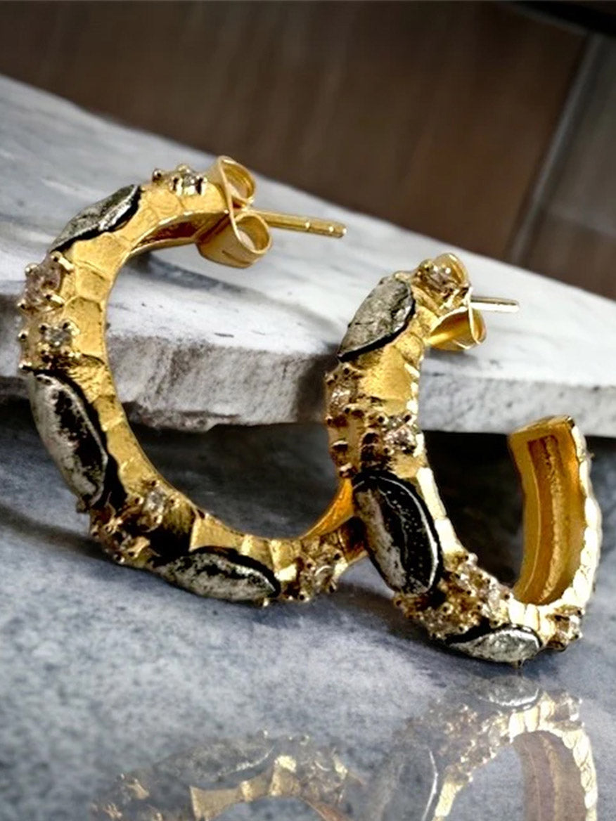 The Tat2 Designs Alma Hoops 1" in Gold & Vintage Silver feature textured gold with unique metal detailing and embedded silver accents from the Tat2 Prince Collection, shown on a marble surface.