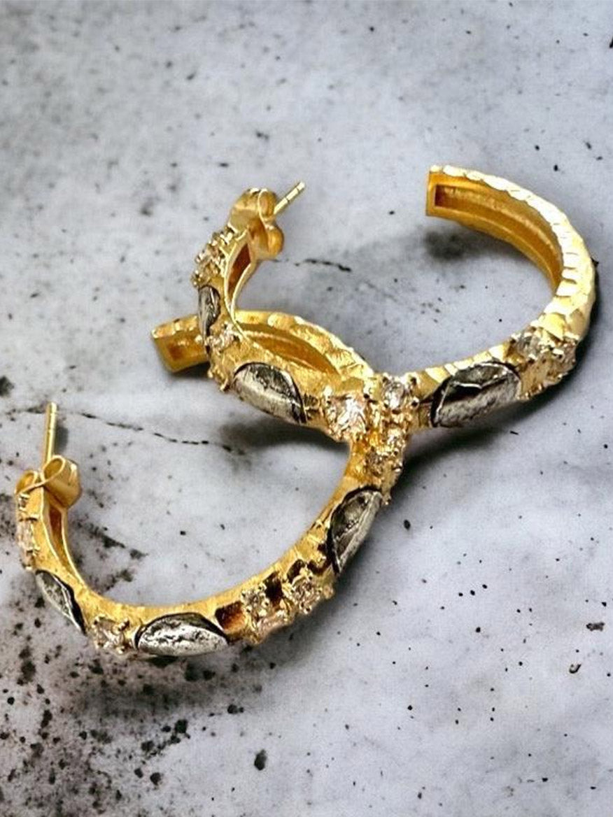 The Tat2 Designs Alma Hoops 1.5" in Gold & Vintage Silver feature gold hoops with vintage silver leaf-shaped accents and clear stones, shown on a marble surface.
