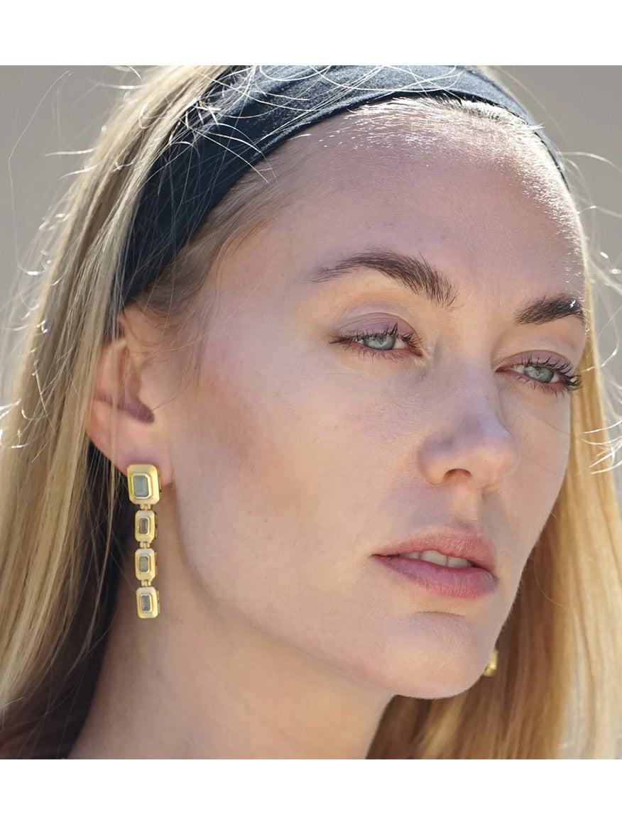 A woman with long blonde hair wears a black headband and Tat2 Designs Labradorite Beryl Earrings in Gold, effortlessly blending vintage and modern trends as she gazes slightly away from the camera.