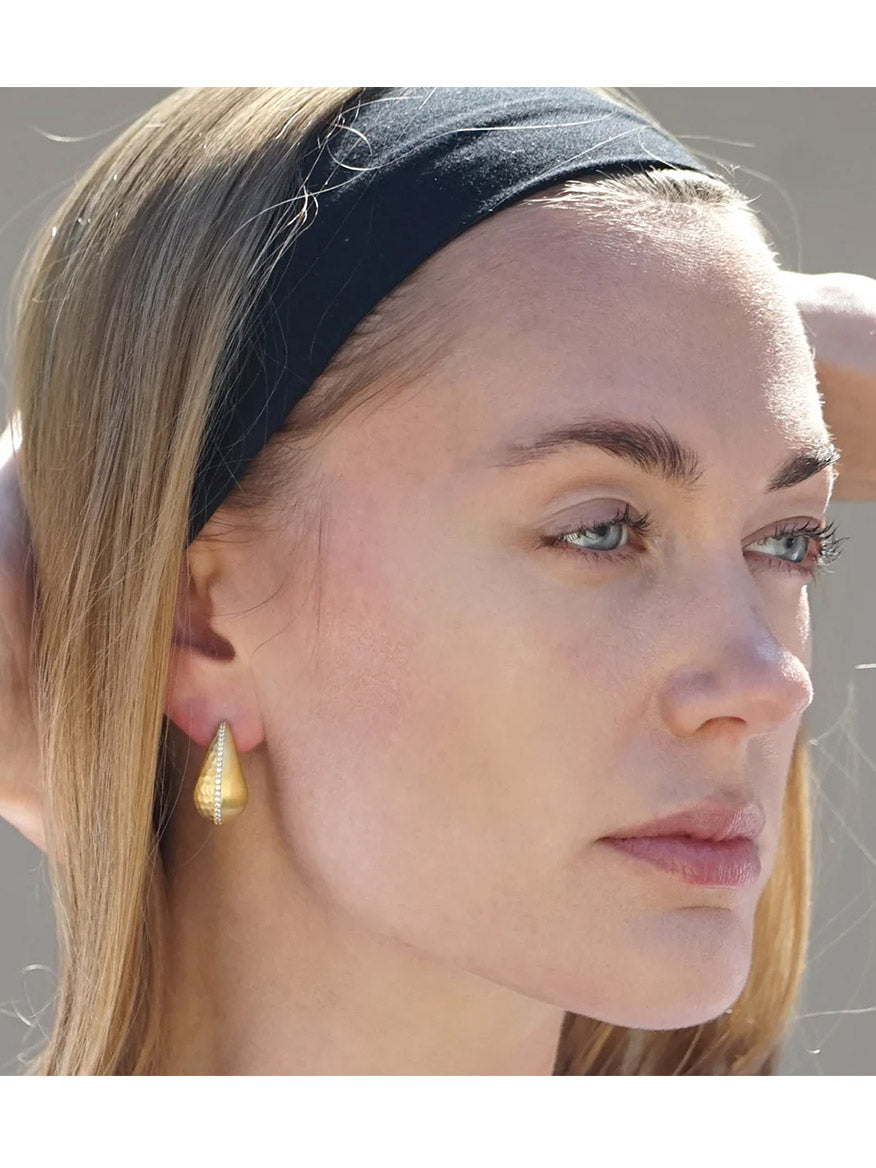 A woman with light eyes and light brown hair wears a black headband and Tat2 Designs Gia Teardrop Earrings in Gold, gazing into the distance.