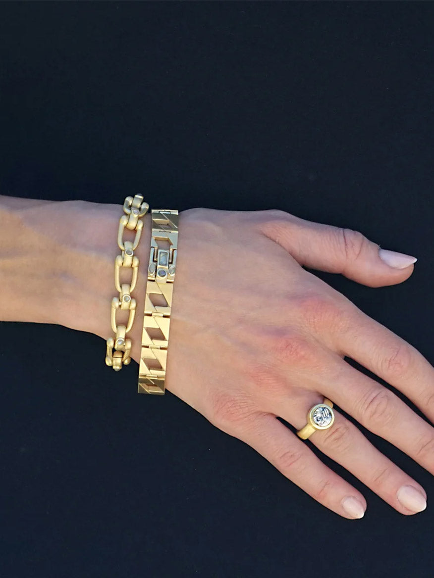A hand with the Tat2 Designs Labradorite Piatta Chain Bracelet in Gold and a gold ring with a round clear stone against a black background.
