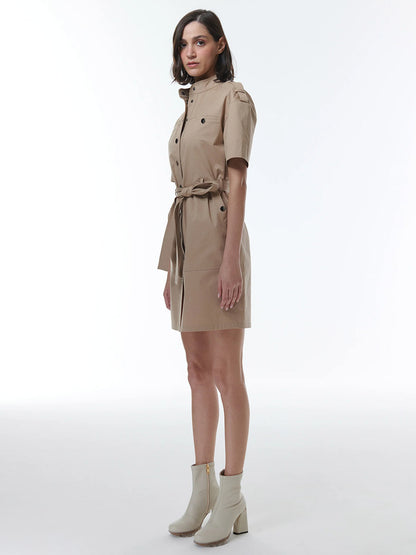 A woman wearing a beige THEO The Label Thallo Safari Dress in Sand with a flattering silhouette and white ankle boots stands against a plain background.