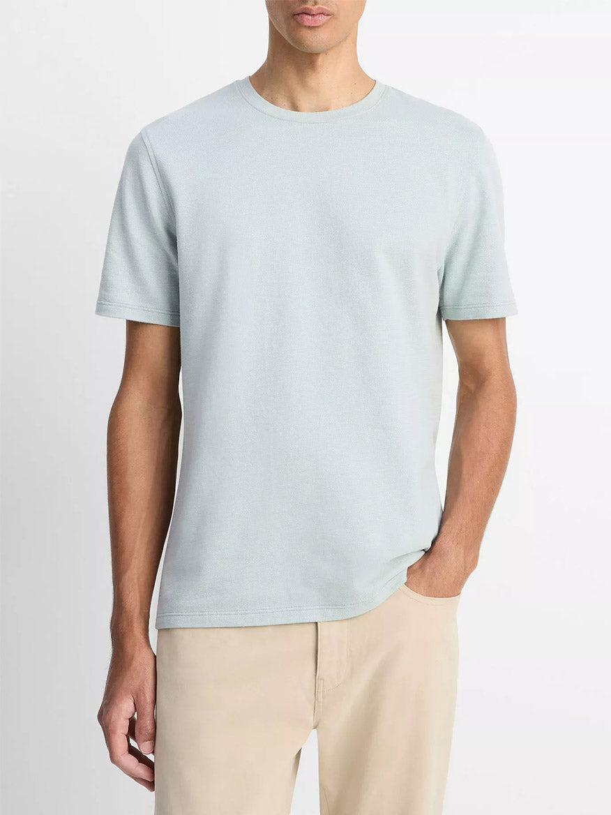 A man wears the Vince Birdseye Jacquard Pima Cotton T-Shirt in Skylight with beige pants, standing against a plain background with one hand in his pocket.