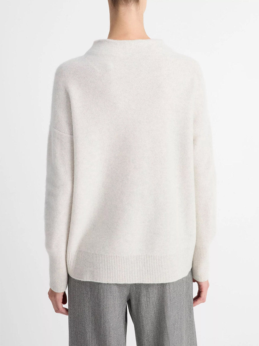 A person stands facing away from the camera, wearing a loose-fitting Vince Plush Cashmere Funnel-Neck Sweater in Heather Platinum and gray trousers against a plain white background.