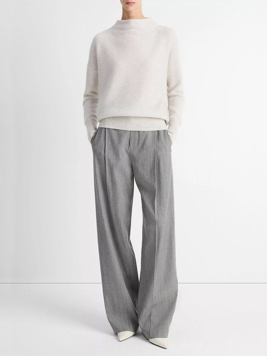 A person wears the Vince Plush Cashmere Funnel-Neck Sweater in Heather Platinum, paired with gray wide-leg trousers and white pointed shoes, standing against a plain white background with hands in pockets.