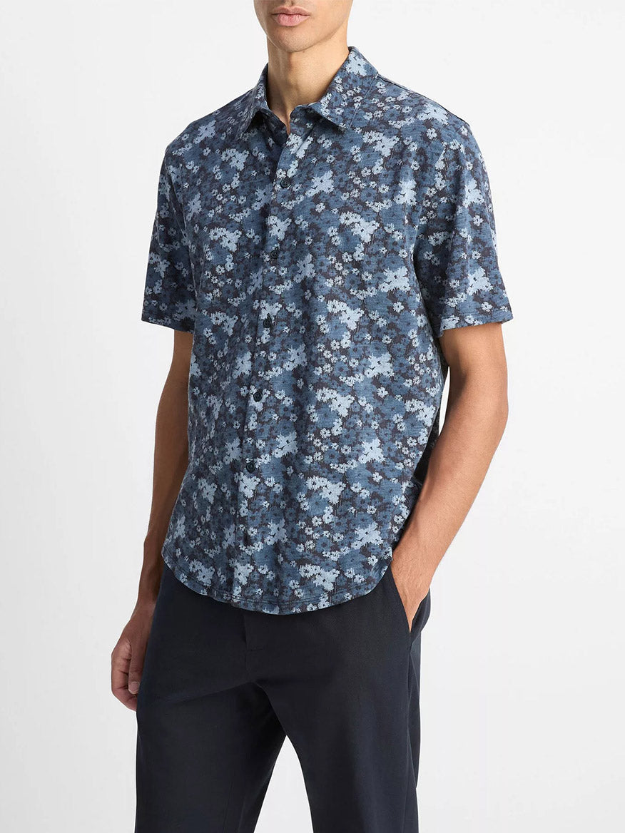A person wearing the Vince Floral Slub Cotton Short-Sleeve Button-Front Shirt in Coastal Blue with dark pants, standing with one hand in their pocket.