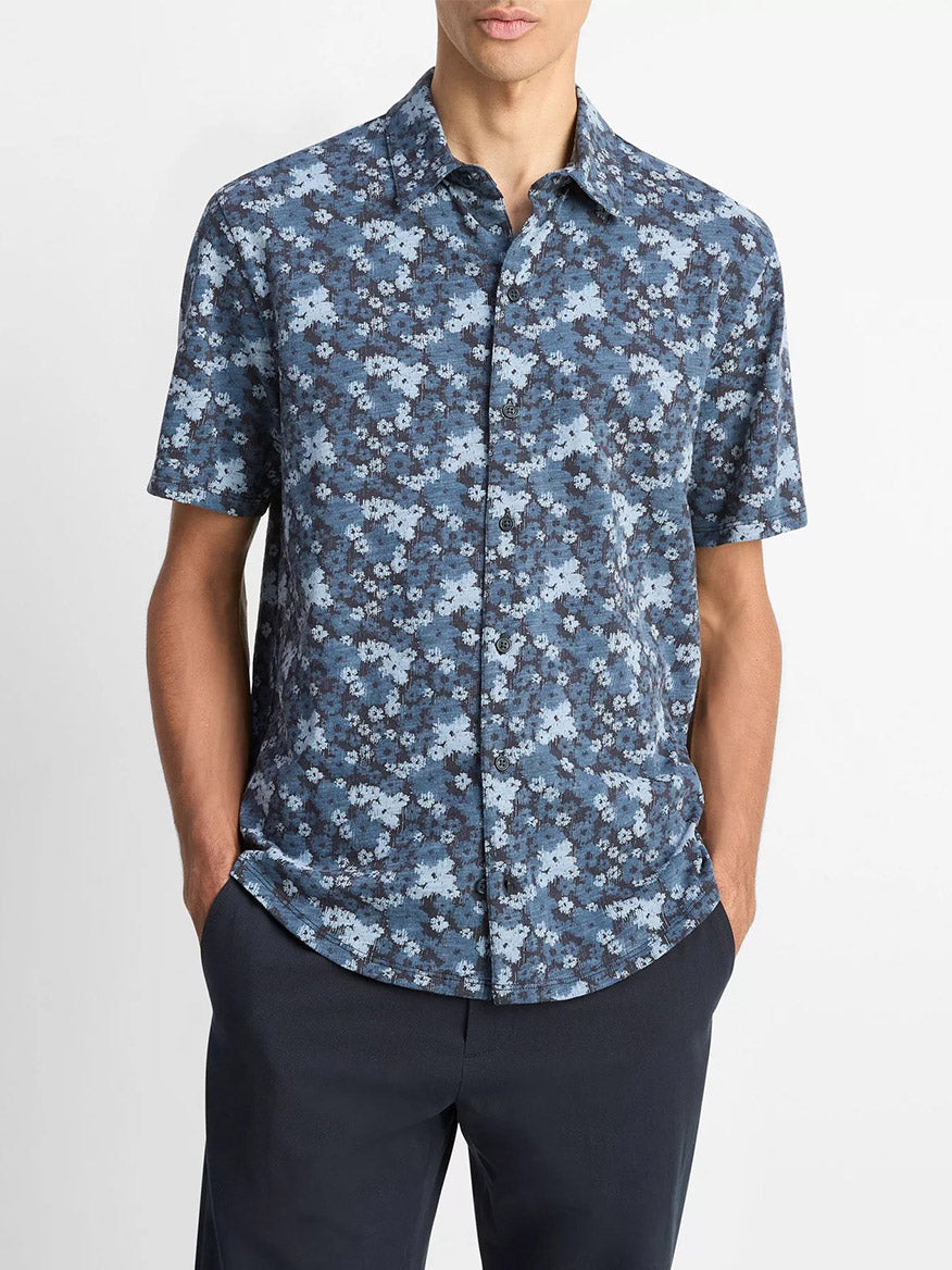 A person models the Vince Floral Slub Cotton Short-Sleeve Button-Front Shirt in Coastal Blue with dark pants, hands in pockets, standing against a plain white background.