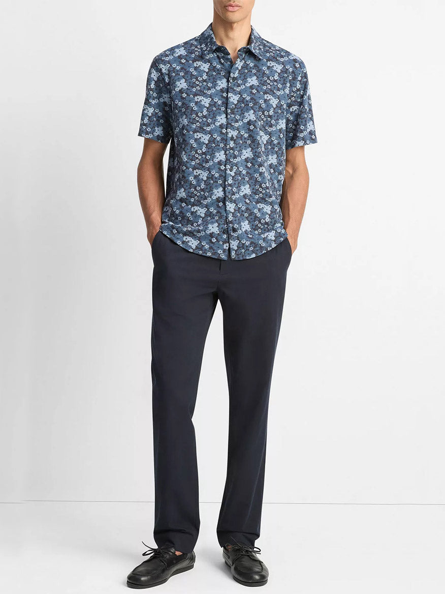 A man stands against a white background, wearing the Vince Floral Slub Cotton Short-Sleeve Button-Front Shirt in Coastal Blue, paired with dark trousers and black shoes, hands in pockets.