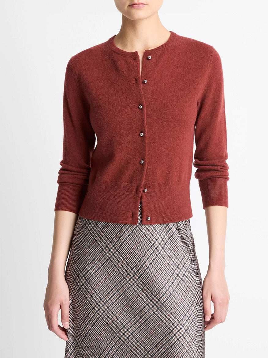 A woman wears the Vince Metal-Button Wool & Cashmere-Blend Cardigan in Siena, paired with a high-waisted plaid skirt, standing against a plain white background.