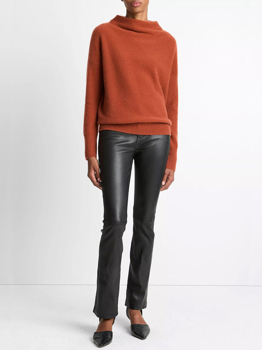 A person wearing the Vince Plush Cashmere Funnel-Neck Sweater in Tuscan Red, black faux leather pants, and black pointed shoes stands against a plain white background.