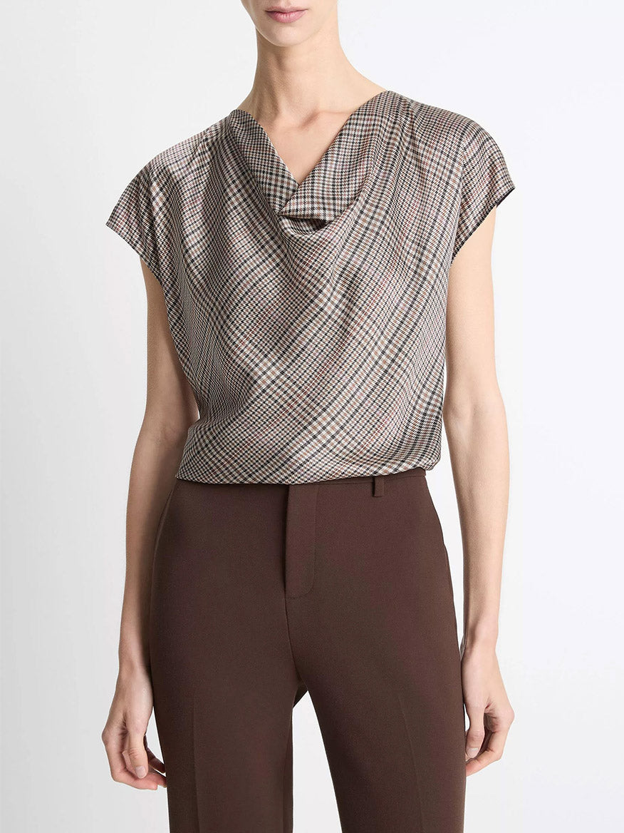 A person wears a Vince Pressed Plaid Silk Cowl-Neck Blouse in Multi Plaid, short-sleeved and tucked into high-waisted brown pants, shown from shoulders to hips against a plain background.