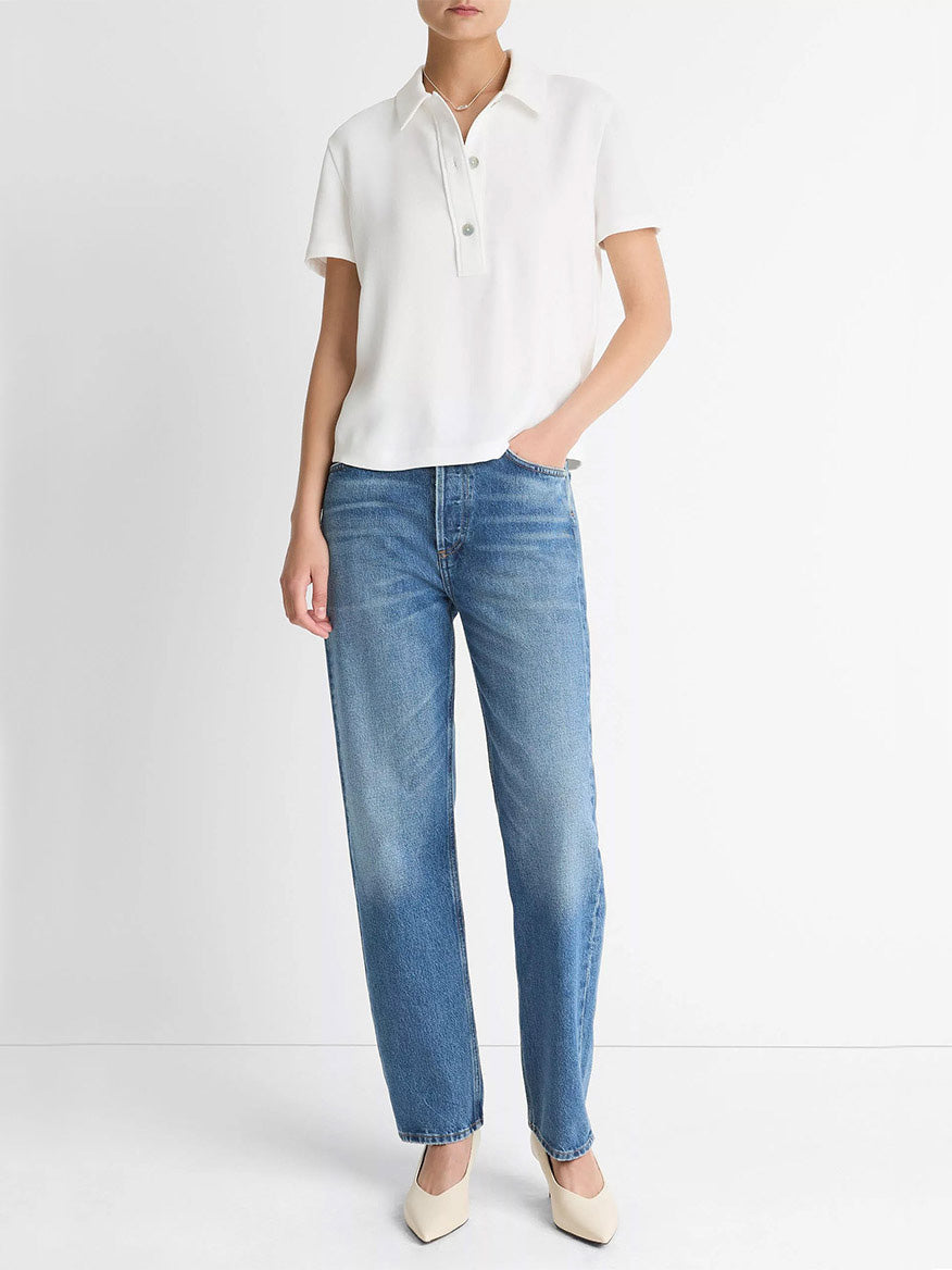A person stands against a white wall wearing the Vince Crepe Polo Shirt in Off-White with agoya shell buttons, blue straight-leg jeans, and beige pointed-toe shoes.