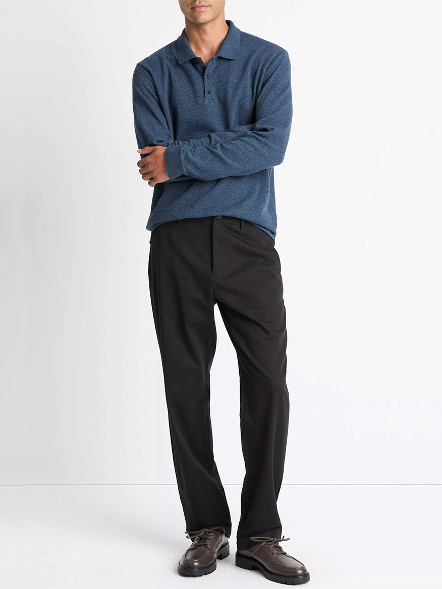 A man stands against a white background wearing the Vince Heathered Double-Face Cotton-Blend Polo Shirt in Heather Coastal Blue/Heather Fog, paired with black trousers and brown boat shoes.