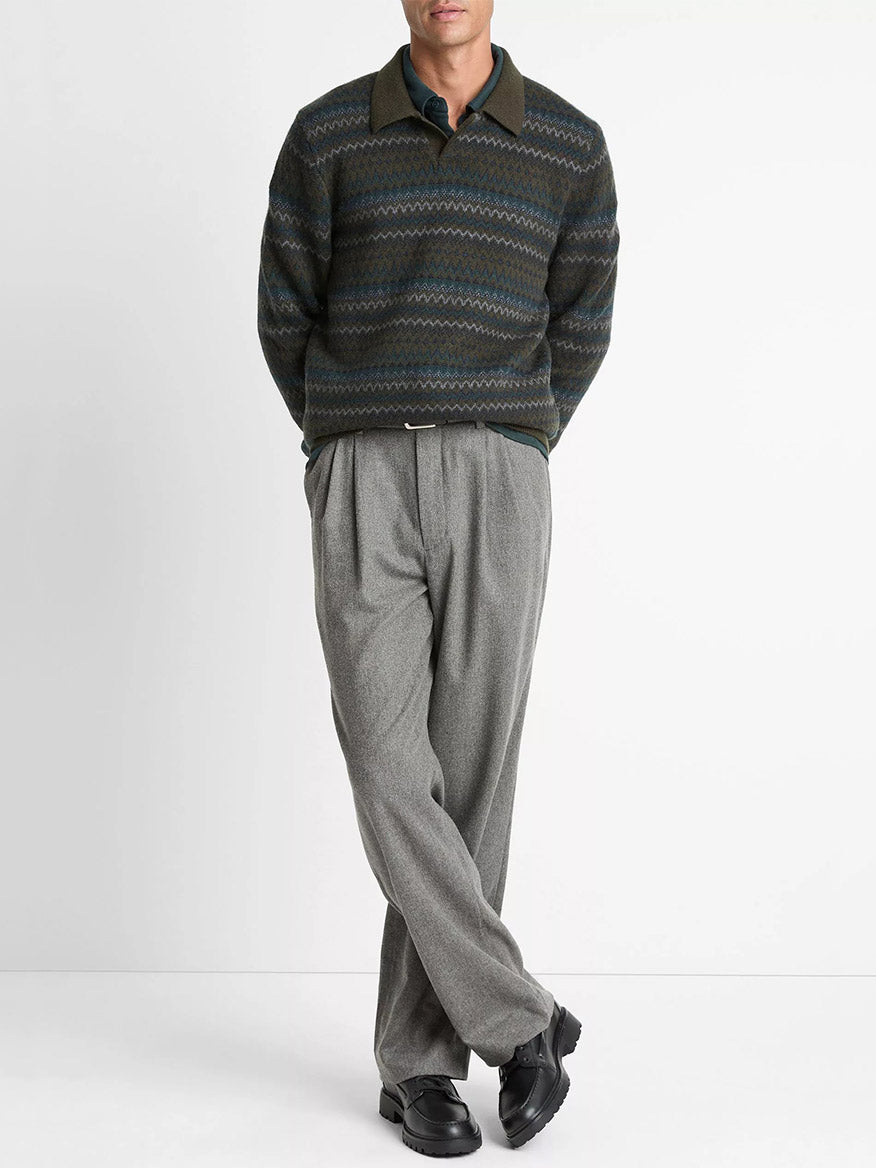 A person stands against a white background wearing the Vince Fair Isle Wool-Cashmere Johnny-Collar Sweater in Dark Leaf Combo, light grey pleated trousers, and black shoes, arms relaxed and legs crossed.