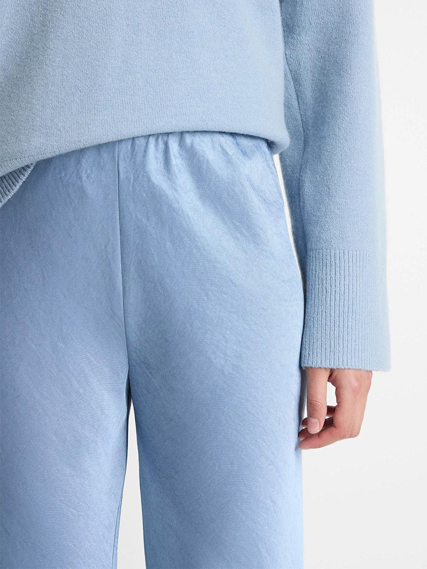 A person wearing Vince Fluid Satin Bias Pant in Pacific, high-waisted with a wide leg fit, shown from the waist down with one hand visible.