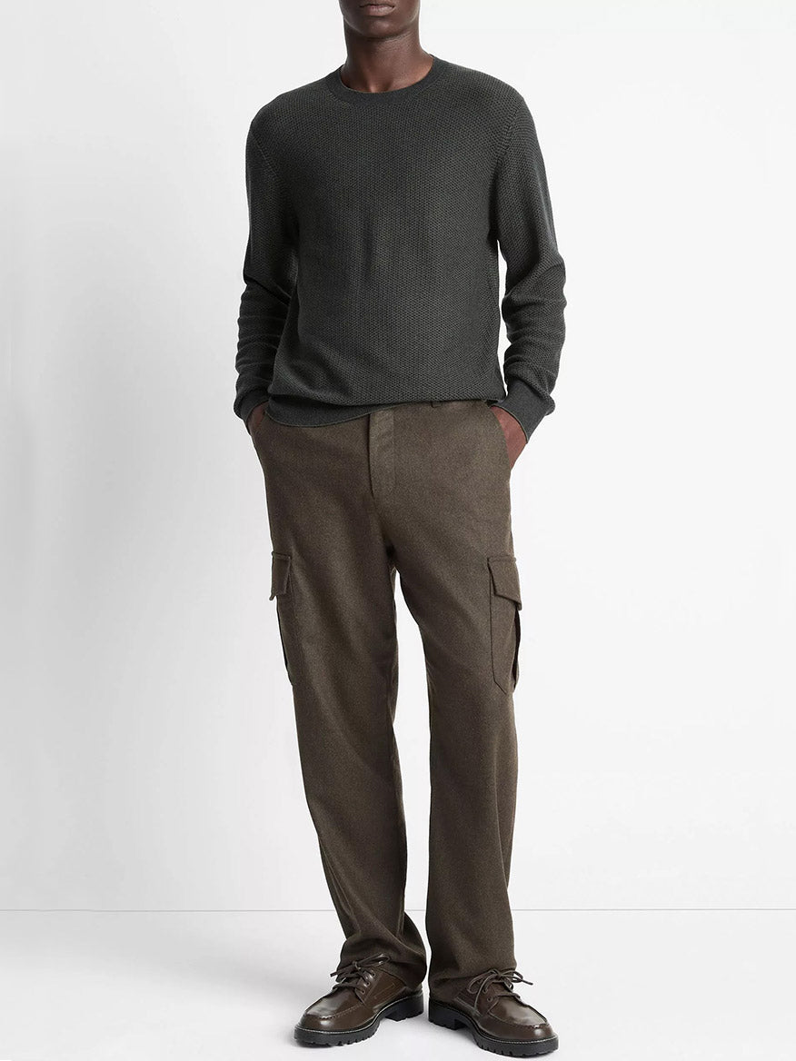 A person stands against a plain background wearing the Vince Geo-Jacquard Wool-Blend Crew Neck Sweater in Driver Green/Coastal Blue, brown cargo pants, and dark shoes, with hands in their pockets.
