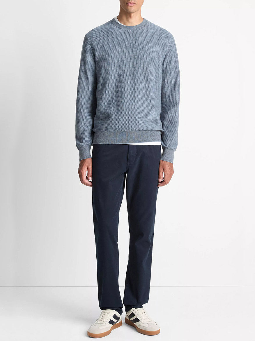A man stands against a plain background, wearing the Vince Geo-Jacquard Wool-Blend Crew Neck Sweater in Parlor Blue/Medium Heather Grey, dark navy pants, and white sneakers with navy accents.