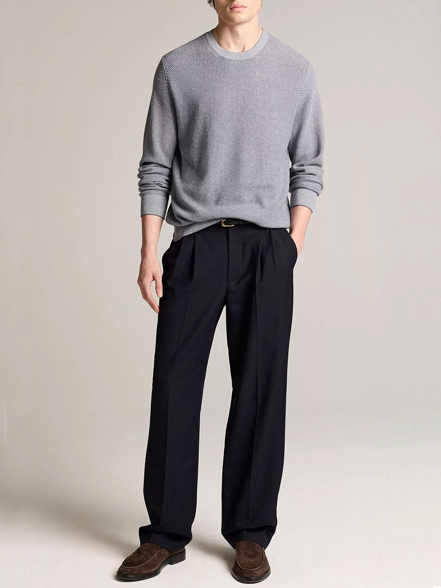 A person stands wearing the Vince Geo-Jacquard Wool-Blend Crew Neck Sweater in Late Evening/Classic Cream, paired with black pleated trousers, a black belt, and brown loafers against a plain light background.