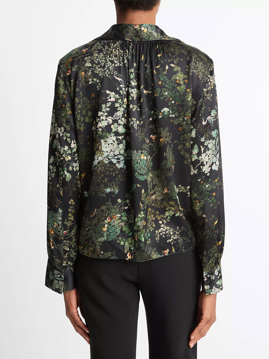 A person is seen from the back wearing the Vince Smocked Grape Vine Silk Long-Sleeve Blouse in Black Sky, featuring a green and white grape vine print, paired with black pants.