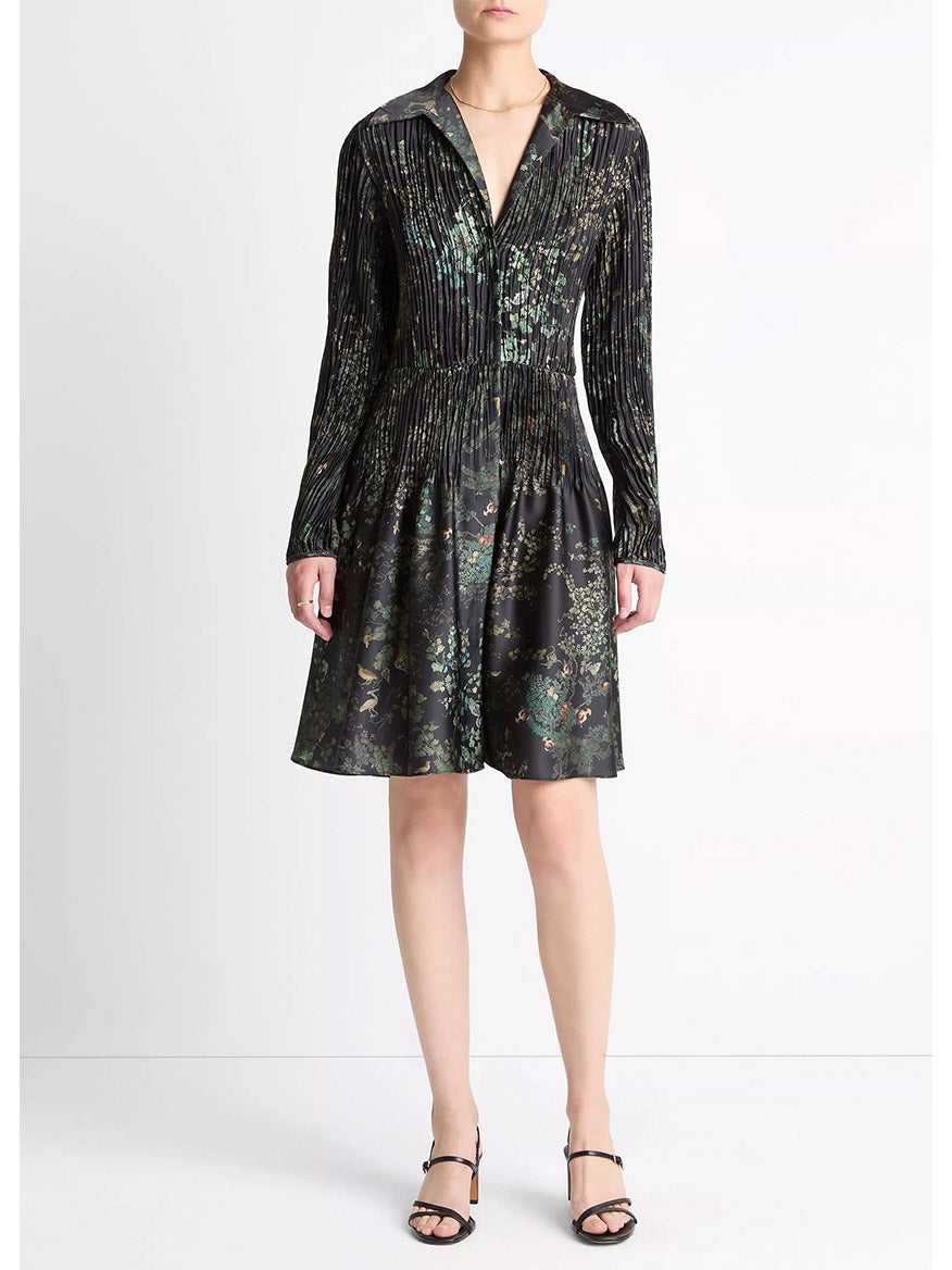 A woman wears the Vince Grape Vine Crushed Satin Shirt Dress in Black Sky, featuring long sleeves, an elasticized waist, and a pleated skirt, paired with black sandals against a plain white background.