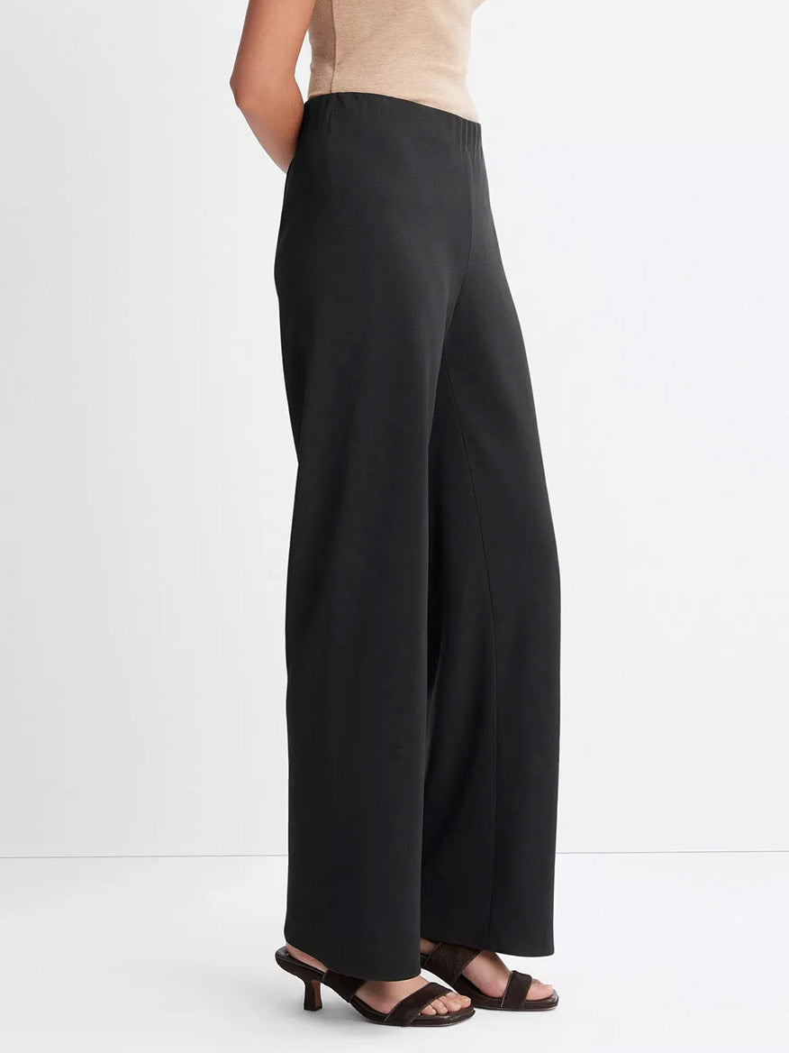 Someone stands against a plain white background wearing the Vince High-Waist Crepe Bias Pant in Black with wide legs and a hidden elastic waistband, paired with a beige sleeveless top and black heeled sandals.