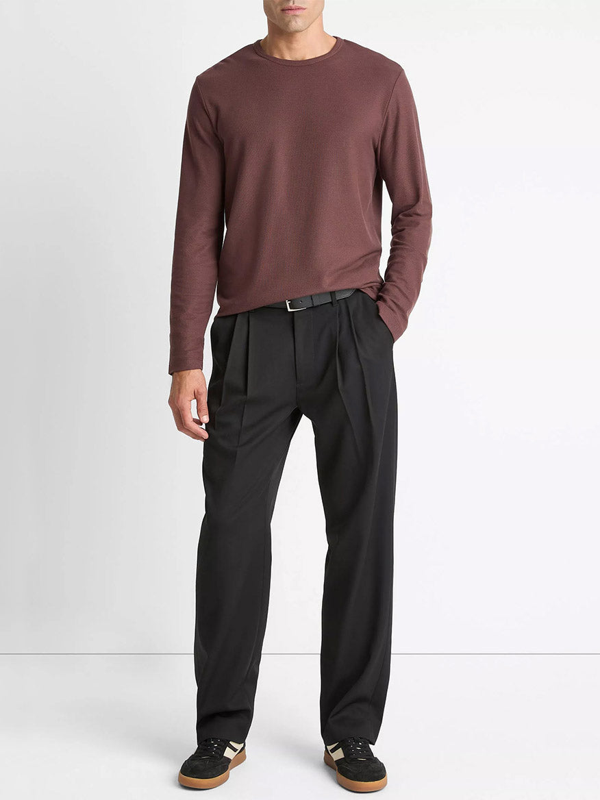 A man stands against a white background wearing a Vince Pima Cotton Piqué Long-Sleeve T-Shirt in Washed Port, black pleated trousers with a belt, and black sneakers with gum soles.