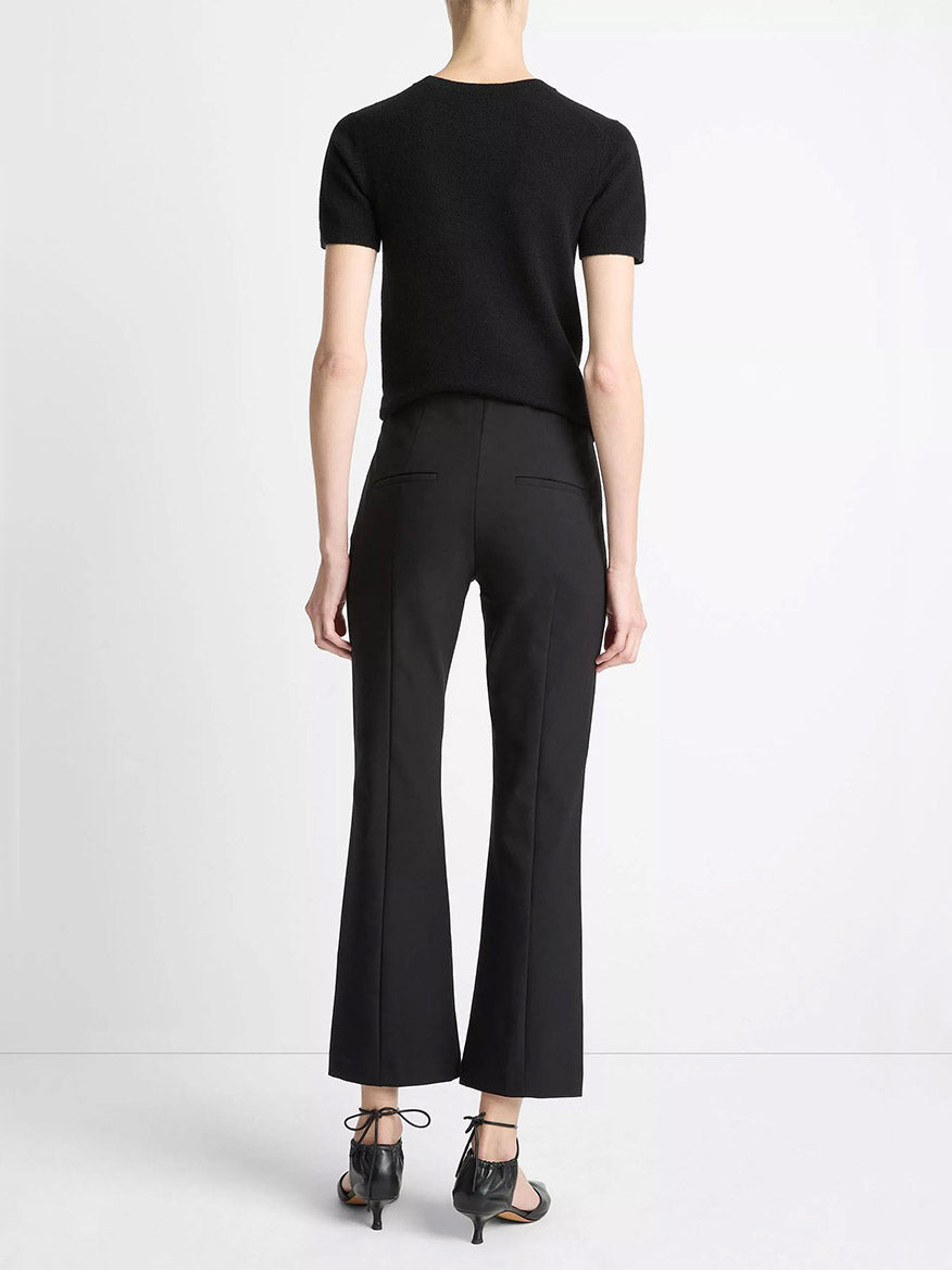 A person stands facing away against a white background, wearing a short-sleeve black top, Vince Mid-Rise Pintuck Crop Flare Pant in Black, and black heeled sandals.