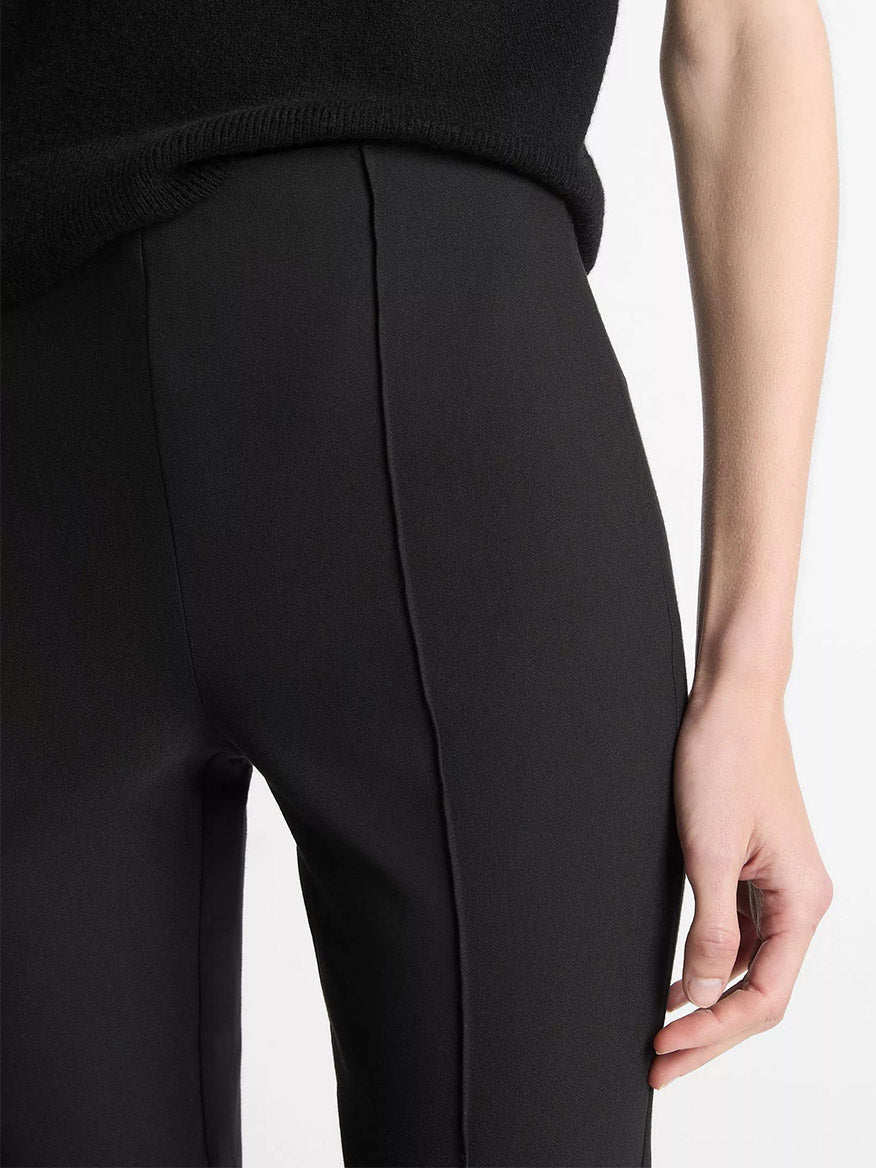 Close-up of a person wearing the Vince Mid-Rise Pintuck Crop Flare Pant in Black with a black top, right arm resting by their side, against a plain background.